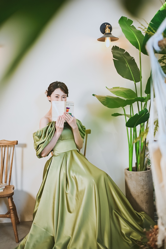 椅子に座り本で口元を隠す、フリルのオリーブグリーンのサテンのカラードレスの花嫁のスタジオフォト