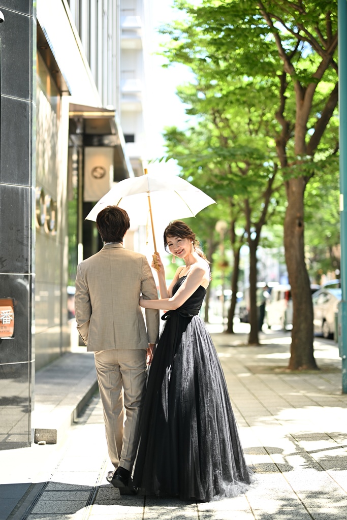 並木道で日傘を差して新郎と腕を組む、ブラックのカラードレスの花嫁の後ろ姿の前撮り