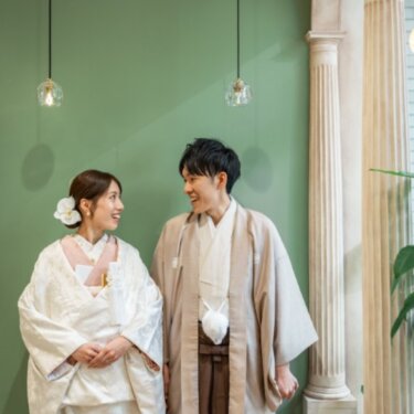 くすみグリーンの壁と円柱のアーチの前で顔を見合わせて笑う、羽織袴の新郎と白無垢の花嫁のスタジオフォト