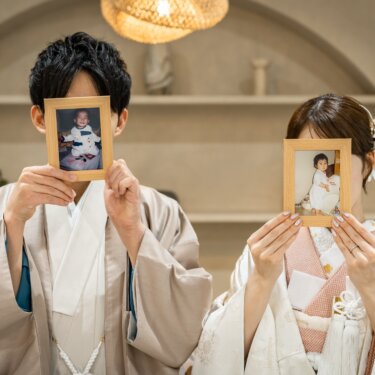 幼い頃の写真を入れたフォトフレームで顔を隠す、羽織の新郎と白無垢の花嫁。成長を感じる和装前撮りのアイテム