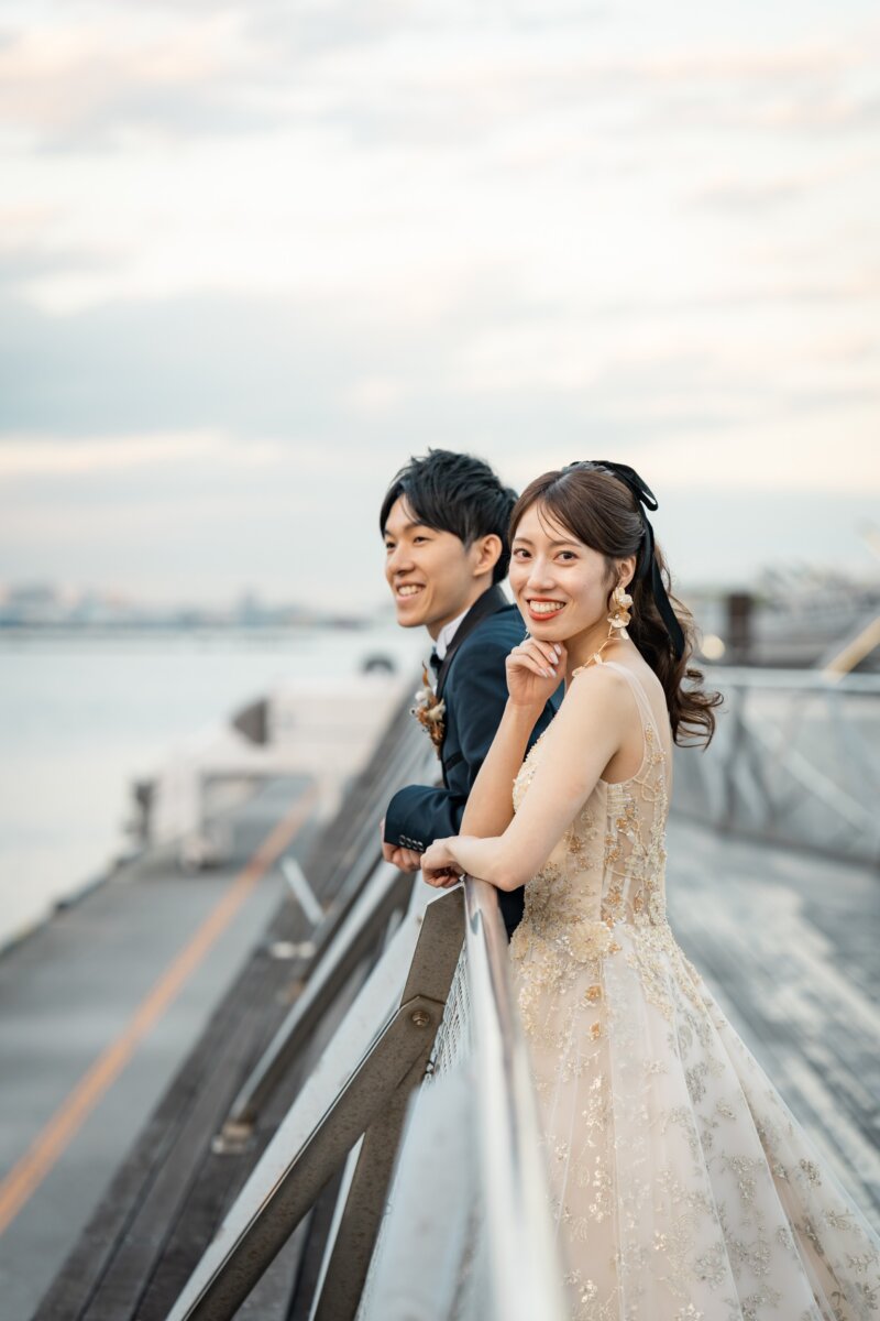 大さん橋の手すりにもたれてカメラに微笑む、ネイビーのタキシードの新郎とカラードレスの花嫁の前撮り