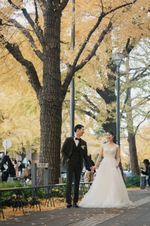 日本大通りの黄色いイチョウ並木の歩道を、手をつないで歩く新郎新婦を引きで写した秋のフォトウェディング