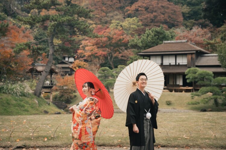 三渓園の古民家と紅葉の山を背景に、赤と白の和傘を差す色打掛の花嫁と紋付袴の新郎の秋の前撮り