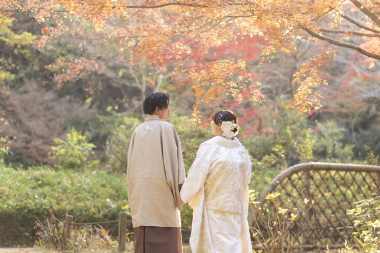 紅葉の木々を見上げながら手をつなぐ、羽織袴の新郎と白無垢の花嫁の後ろ姿。久良岐公園の秋の和装前撮り