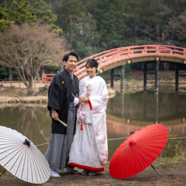 称名寺の反橋が水面に映る池のほとりで、紅白の和傘を置いて並ぶ新郎新婦。水鏡が美しい和装ロケーションフォト