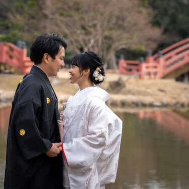 朱色の反橋を背に、手を取り見つめ合って笑う新郎新婦。称名寺の庭園で撮る和装前撮り