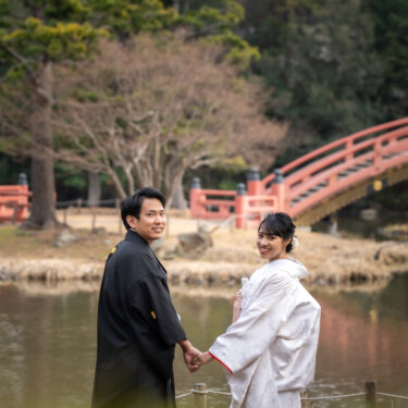 称名寺の池のほとりで手をつなぎ、振り返ってほほえむ新郎新婦。朱色の反橋が映える結婚写真