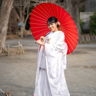 白梅が咲き始めた境内で、赤い和傘を掲げてほほえむ白無垢の花嫁のポートレート。春の和装フォトウェディング