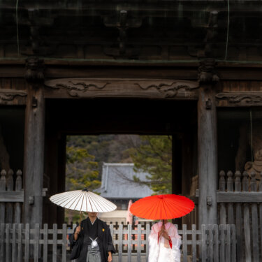 称名寺の仁王門の下で、白と赤の和傘を差して離れて立つ新郎新婦の和装ロケーションフォト