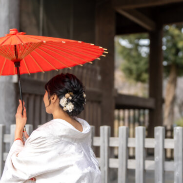 称名寺の仁王門の柱越しに、赤い和傘を差す白無垢の花嫁の後ろ姿。花の髪飾りが映える和装フォトウェディング