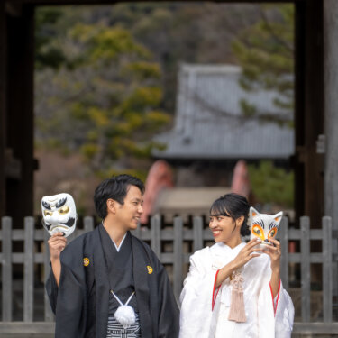 称名寺の仁王門の下で、天狗と狐のお面を掲げておどける新郎新婦。遊び心あるお面の和装前撮り