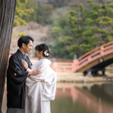 大木と朱色の反橋を背景に、額を合わせて寄り添う紋付袴の新郎と白無垢の花嫁の横顔の結婚写真
