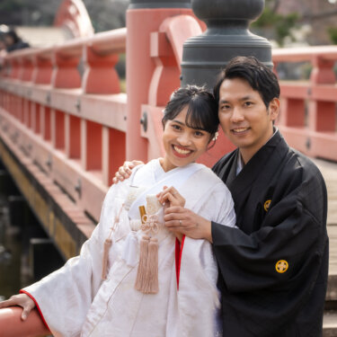 朱色の欄干のそばで頬を寄せ、カメラに笑う白無垢の花嫁と紋付袴の新郎の和装フォトウェディング