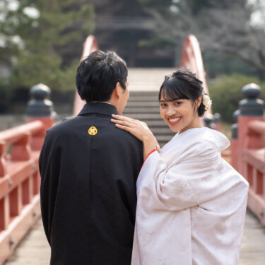 朱色の反橋の上で新郎の肩に手を添え、振り返って笑う白無垢の花嫁と後ろ姿の新郎の和装前撮り