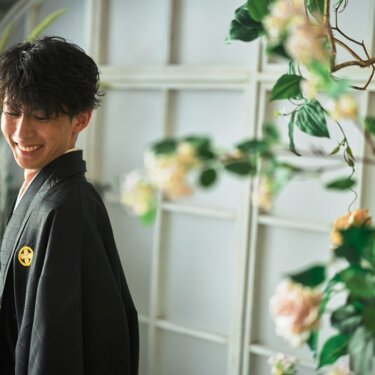花とグリーンを前景に、うつむいてほほえむ黒紋付の新郎のポートレート。和装スタジオフォト