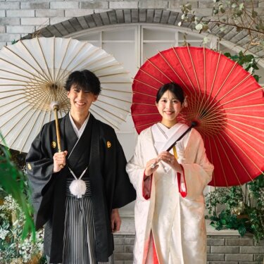 花の装飾のアーチ窓の前で、白と赤の和傘を掲げてほほえむ紋付袴の新郎と白無垢の花嫁の和装前撮り