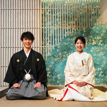 青緑の花柄の壁の前で並んで正座し、満面の笑みを見せる紋付袴の新郎と白無垢の花嫁の結婚写真