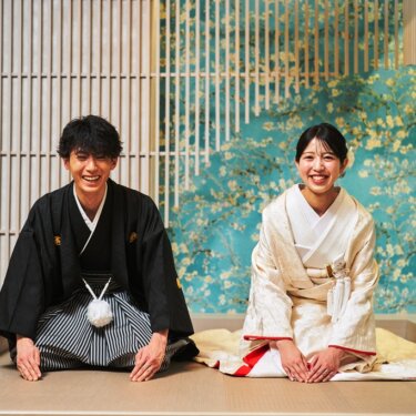 青い花柄の壁のセットに正座し、声を上げて笑う紋付袴の新郎と白無垢の花嫁の和装スタジオフォト