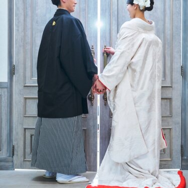 光の差す大きな扉の前で手をつなぎ、振り返る紋付袴の新郎と白無垢の花嫁の後ろ姿の和装前撮り