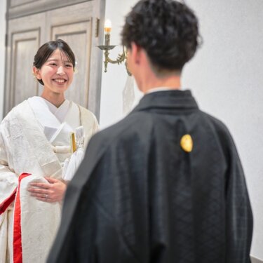 新郎の後ろ姿越しに、懐剣と末広を差した白無垢の花嫁が笑顔を見せるファーストミートの和装フォトウェディング