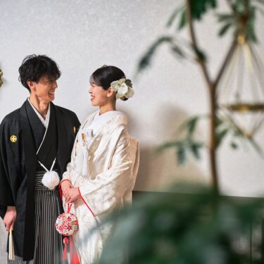観葉植物越しに見つめ合う紋付袴の新郎と手毬を持つ白無垢の花嫁。やわらかな雰囲気の和装スタジオフォト