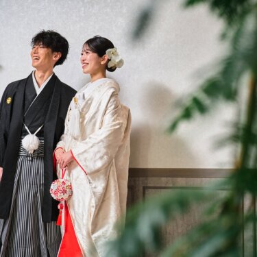 壁灯のある白い壁の前で声を上げて笑い合う紋付袴の新郎と白無垢の花嫁を、観葉植物越しに写した和装フォトウェディング