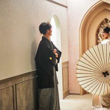 尖塔アーチの前で大きな和傘を広げて振り返る白無垢の花嫁と、壁にもたれる紋付袴の新郎の和装前撮り