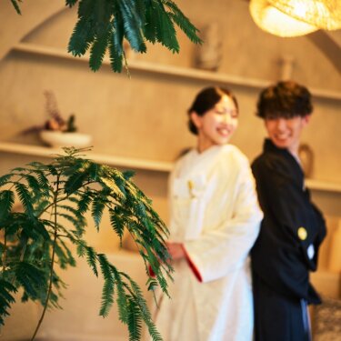 観葉植物越しに、アーチの棚の前で背中合わせに立つ白無垢の花嫁と紋付袴の新郎をぼかして写したやわらかな和装前撮り