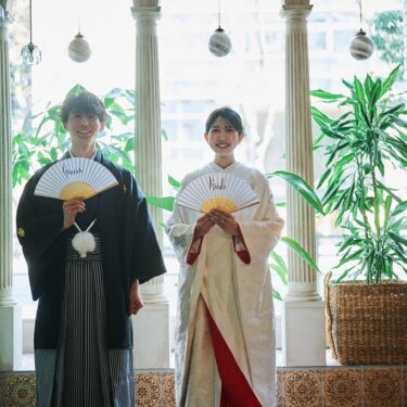 円柱と観葉植物が並ぶ明るい窓辺で「Groom」「Bride」の扇子を掲げる和装前撮り
