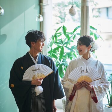 アーチ窓とグリーンを背景に、'Groom''Bride'の扇子を手に見つめ合う紋付袴の新郎と白無垢の花嫁の和装前撮り