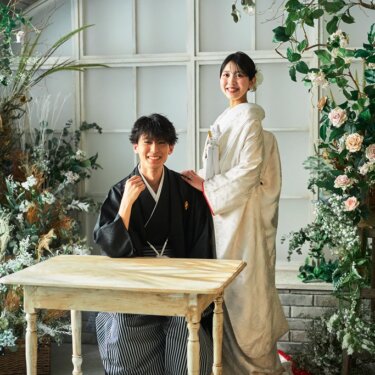 木のテーブルに座る紋付袴の新郎と、肩に手を添えてほほえむ白無垢の花嫁。花のアーチ窓のセットの和装前撮り