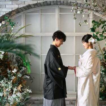 花とドライフラワーのアーチ窓の前で向かい合い、手を取る新郎新婦。スタジオで撮る和装前撮り