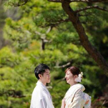 松の枝の下で手を取り見つめ合う、藤色の紋付袴の新郎と淡い色打掛の花嫁。三渓園の和装前撮り
