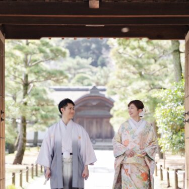 三渓園の門の内側から、参道に立つ淡い色打掛の花嫁と藤色の紋付袴の新郎を写した引きの和装前撮り