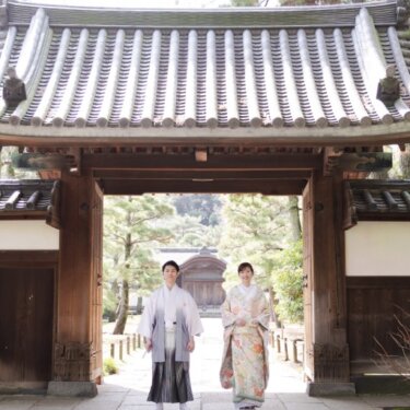 三渓園の瓦屋根の門をくぐって歩いてくる紋付袴の新郎と色打掛の花嫁を、門全体とともに写した和装ロケーションフォト