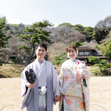 数寄屋造りの建物と松を背に、狐のお面を手にしてカメラに笑う新郎新婦。遊び心あるお面の和装前撮り