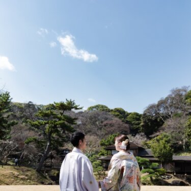 三渓園の茅葺屋根の建物群と青空を望む芝生で、向かい合う新郎新婦の後ろ姿を写した和装前撮り