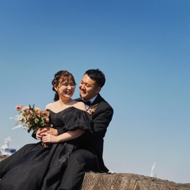 青空の下の岸壁に座り、花嫁を後ろから抱えて笑い合う新郎新婦。ブーケを持った黒いドレスのウェディングフォト