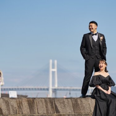 横浜ベイブリッジを望む石積みの岸壁で、立つタキシードの新郎と座って笑う黒いドレスの花嫁の前撮り