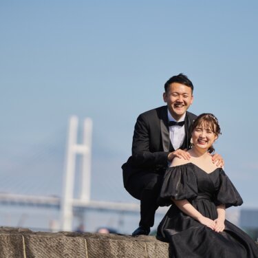 横浜ベイブリッジを背に、座る黒いカラードレスの花嫁の肩に手を添えて大笑いする新郎。港で撮るウェディングフォト