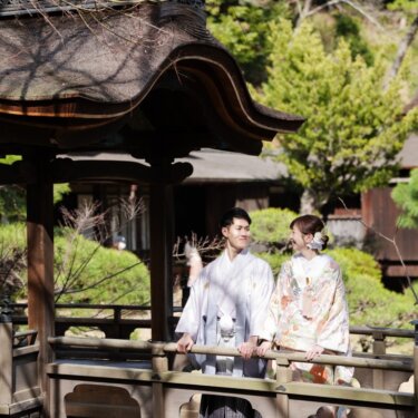 茅葺屋根の建物群を背に、三渓園の屋根付き橋に並ぶ淡い色打掛の花嫁と藤色の紋付袴の新郎の和装前撮り