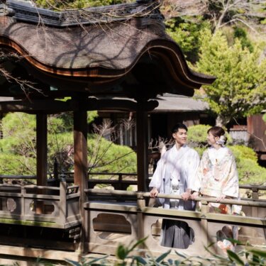 三渓園の唐破風の屋根付き橋の欄干に並ぶ紋付袴の新郎と色打掛の花嫁を、装飾的な梁とともに写した和装前撮り