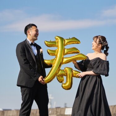 '寿'の文字の金色のバルーンを二人で持ち、大笑いする新郎新婦。青空の下で撮る遊び心ある前撮り