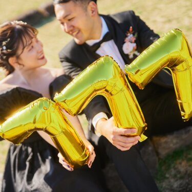 芝生に座り、'777'の金色のバルーンを抱えて笑い合う新郎新婦。黒いドレスとタキシードのウェディングフォト