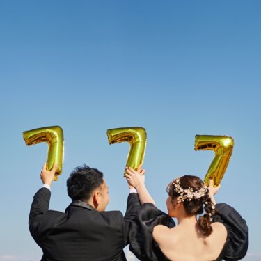 青空に'777'の金色の数字バルーンを掲げる新郎新婦の後ろ姿。小物を使った楽しい前撮り