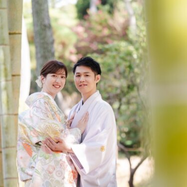 竹の枝を前景にぼかし、寄り添ってカメラにほほえむ淡い色打掛の花嫁と新郎の和装前撮り