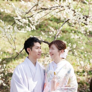 三渓園の白梅の咲く木々を背景に、手を取り見つめ合う淡い色打掛の花嫁と藤色の紋付袴の新郎の前撮り