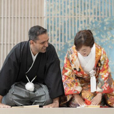 青い花柄の壁と格子のセットで正座し、扇子を前にほほえみ合う新郎新婦の和装フォトウェディング
