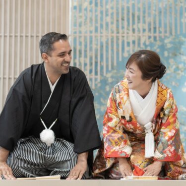 青い花柄の壁の前で正座し、笑い合う新郎と鶴柄の色打掛の花嫁。和モダンなスタジオの和装前撮り