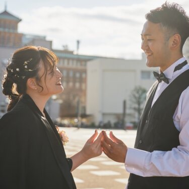 塔のある歴史的な庁舎を背に、指輪を挟んで持ち笑い合う黒いドレスの花嫁とベストの新郎。横浜の街ロケで撮るウェディングフォト
